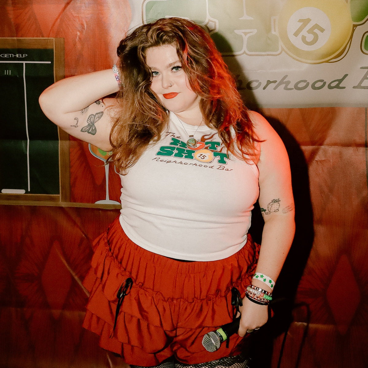 Leanna posing in front of a 'The Hot  Shot Neighborhood Bar' backdrop, wearing The Hot Shot Neighborhood Bar Tank and a red ruffled mini skirt.