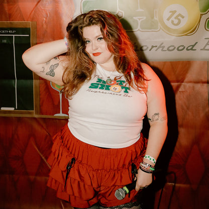 Leanna posing in front of a 'The Hot  Shot Neighborhood Bar' backdrop, wearing The Hot Shot Neighborhood Bar Tank and a red ruffled mini skirt.