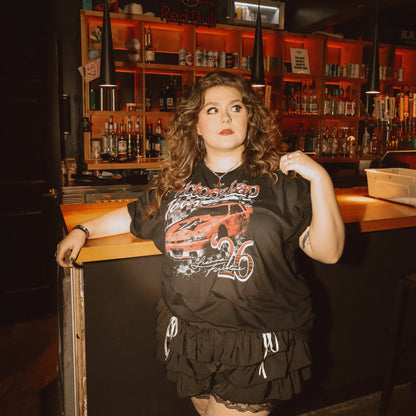 Leanna stands in a dimly lit bar, wearing the Victory Lap Tour t-shirt with black ruffled shorts.